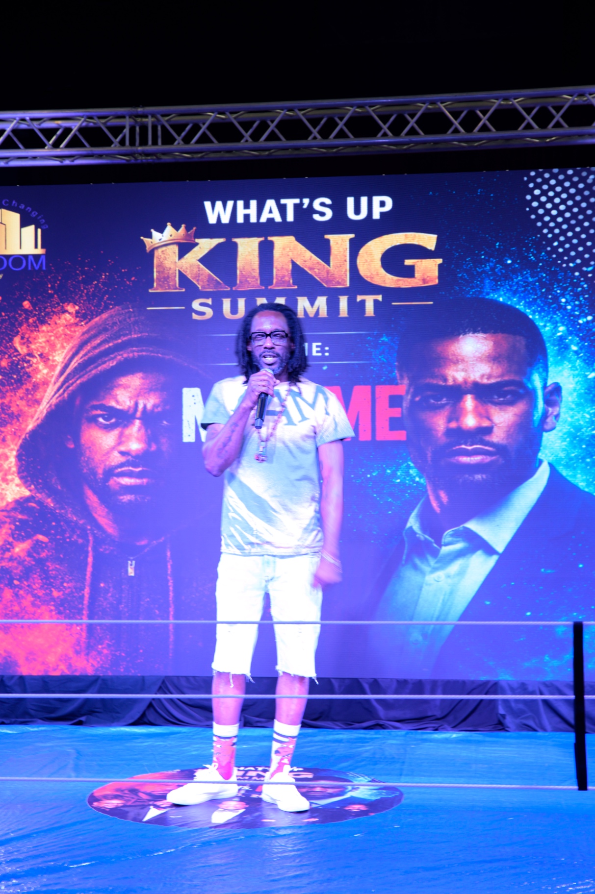 A speaker holding a microphone at center stage before the Me vs. Me summit backdrop